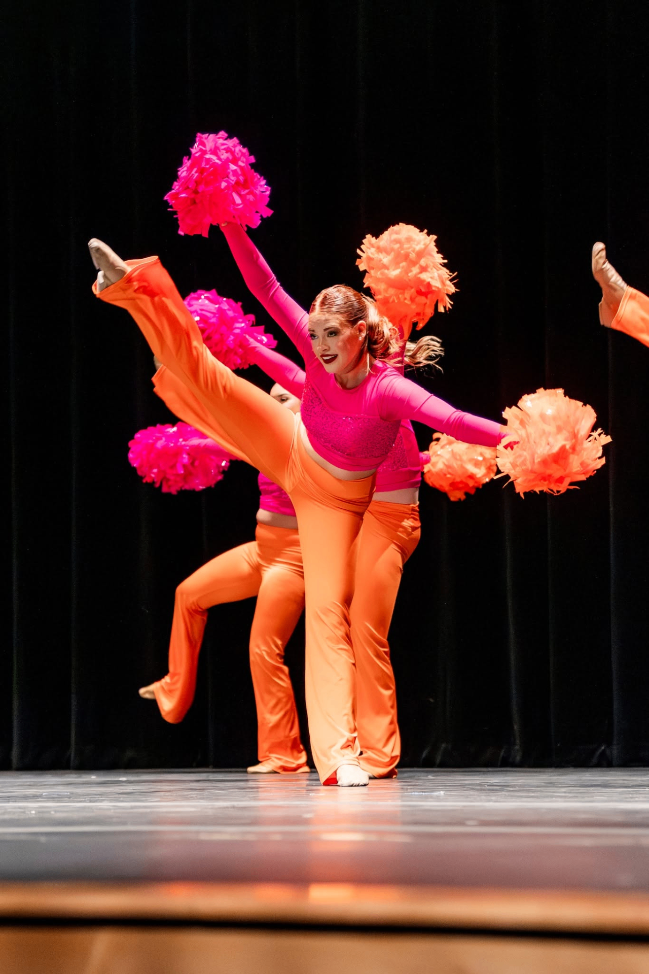 Pom dancers in vibrant pink and orange costumes performing on stage with poms.