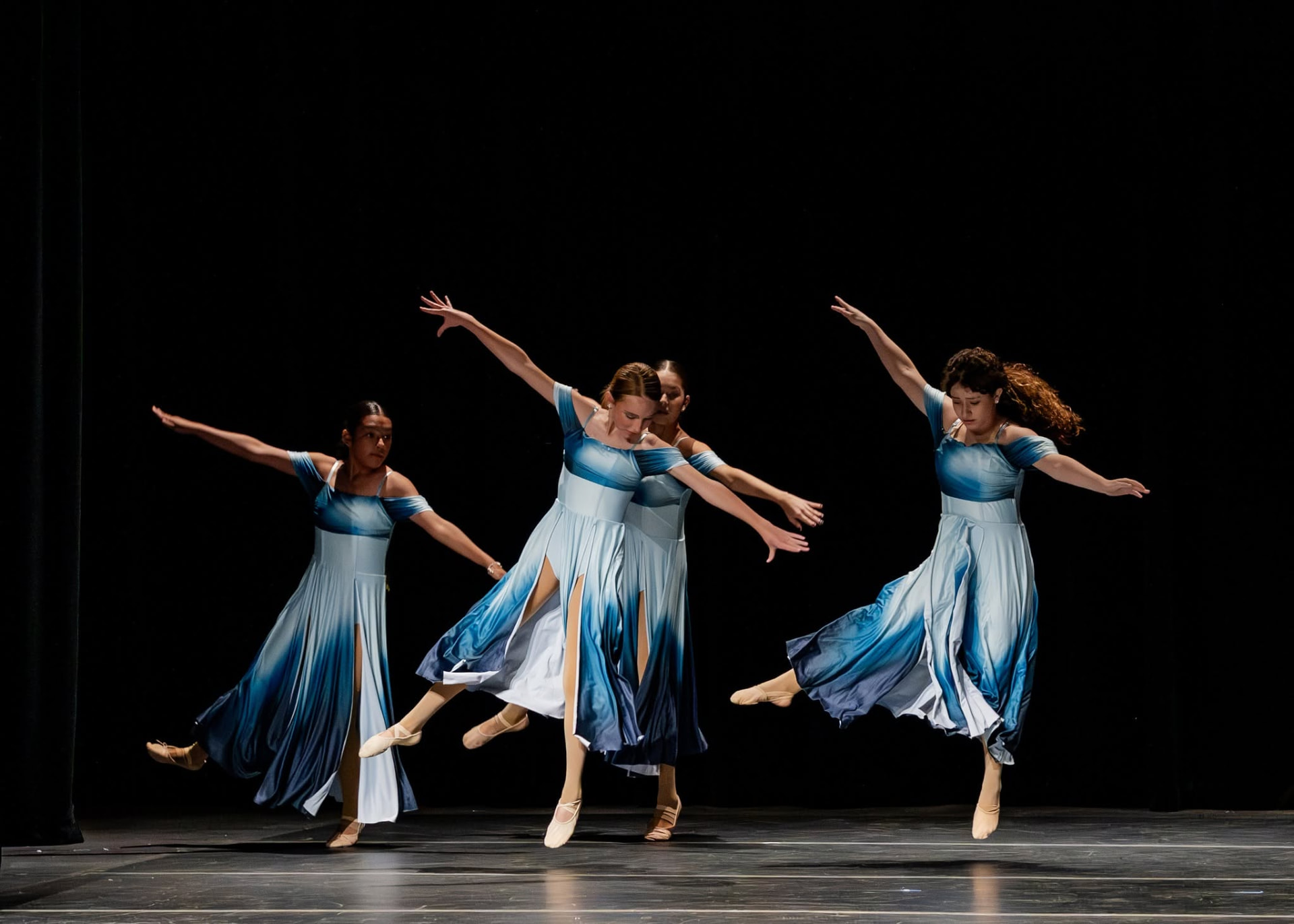 Four dancers in blue ombre dresses leaping across the stage