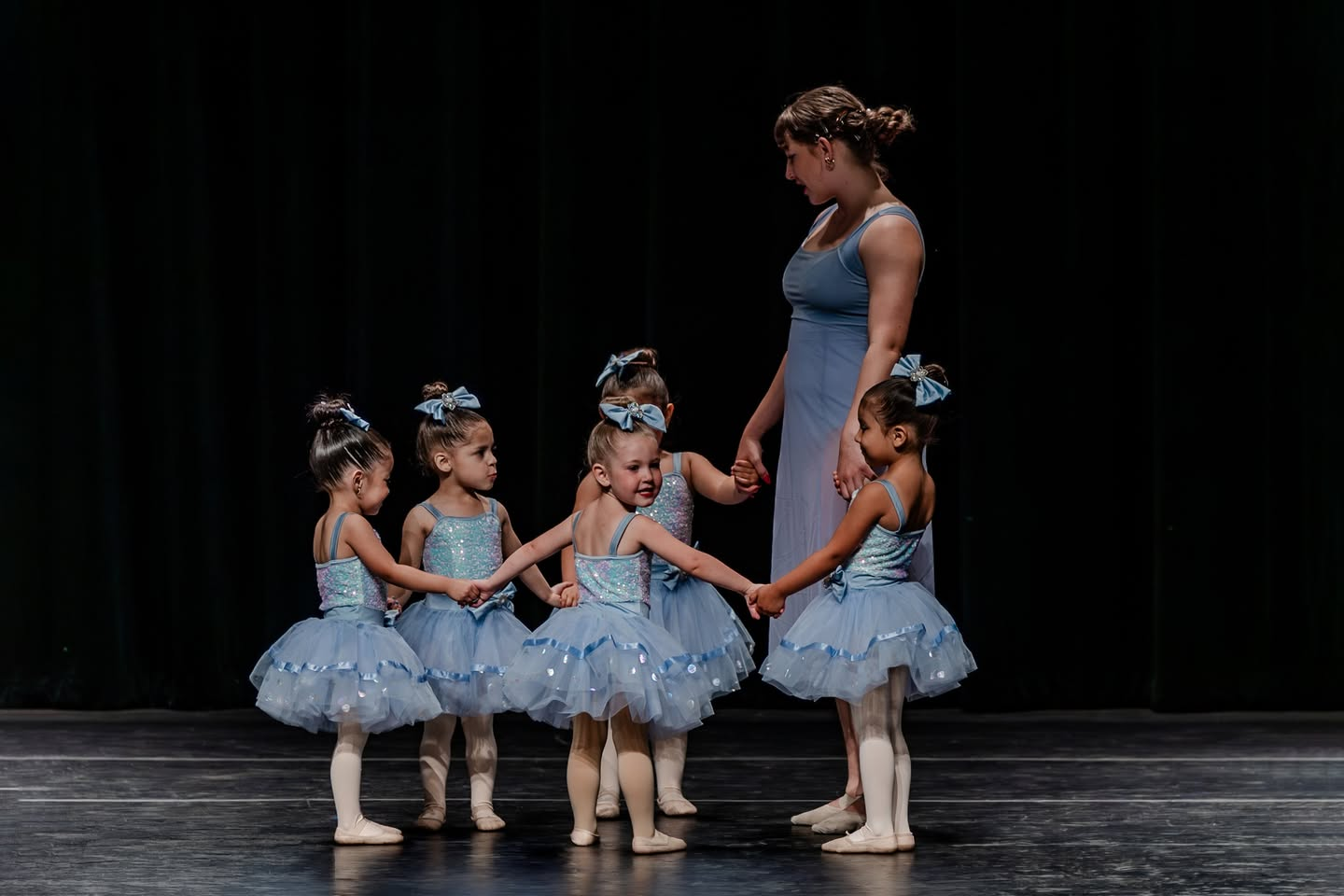 Instructor guiding a circle of young Baby Ballerinas dancers in matching blue tutus on stage.